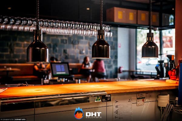 Timber bar counter at History Cafe Essendon under three overhead heat lamps, with wine glasses racked above and the dining room beyond