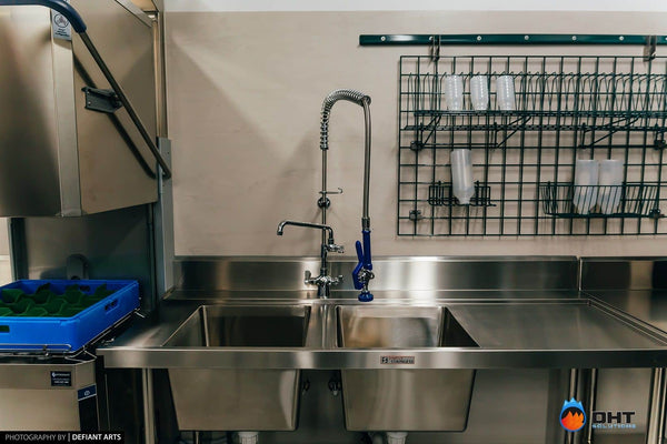 Simply Stainless double sink and pre-rinse spray beside the pass-through dishwasher, with glass racks on the wall