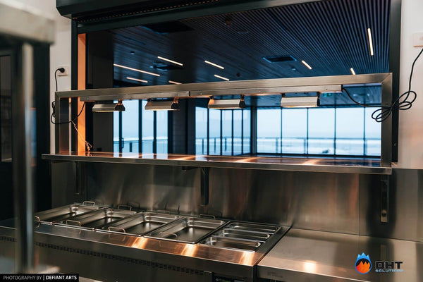 Heat-lamp gantry over the bain marie on the pass, with the ocean visible through the function room windows